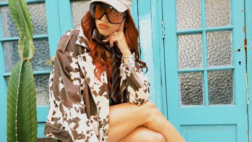 Influencer chilena Lauriturri usando chaqueta con estampado de vaca con fondo colorido celeste, gafas cafés grandes y gorro.