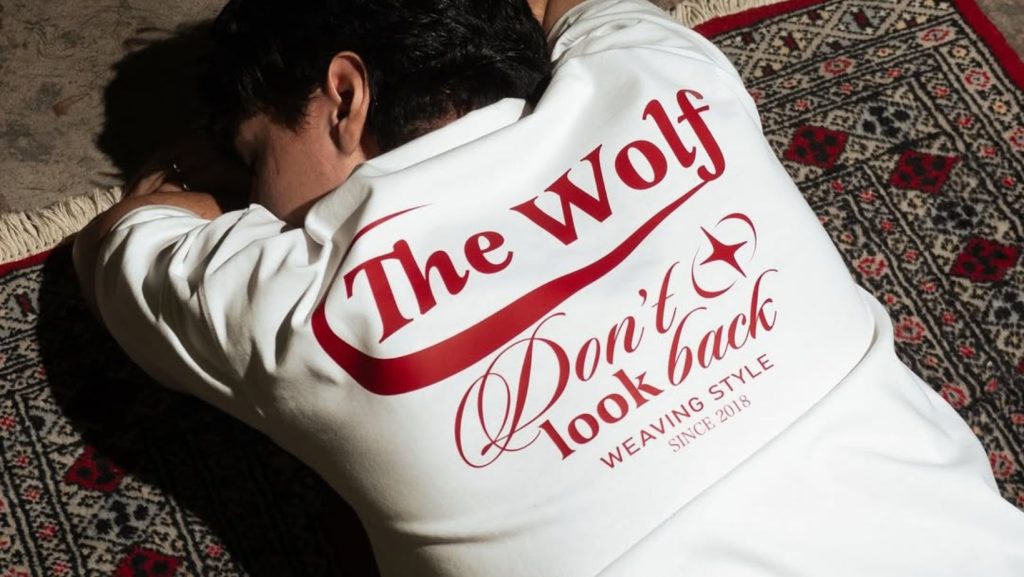 Hombre acostado sobre alfombra usando polera The Wolf, marca chilena. Polera blanca con diseño en rojo. Imagen de The Wolf. Permiso de uso entregado por el dueño de la marca.