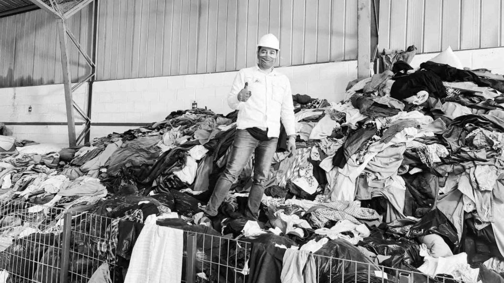 Franklin Zepeda creador de Phyro rodeado de grandes montones de ropa y residuos textiles. Fotografia del Instagram de Procitex Chile.